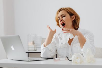 A woman in white yawning while working at a desk, indicating fatigue.