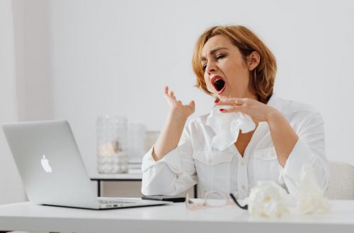 A woman in white yawning while working at a desk, indicating fatigue.