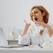 A woman in white yawning while working at a desk, indicating fatigue.